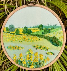 a cross - stitch embroidery project with yellow flowers and green fields in the background is hanging on a tree branch