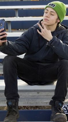 a young man is sitting on a bench and looking at his cell phone while wearing a green beanie