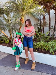 two women standing next to each other in front of palm trees and bushes, one holding a heart shaped balloon