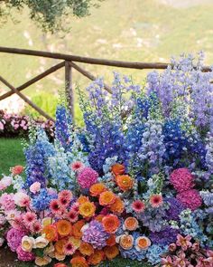 an arrangement of colorful flowers in front of a wooden fence with grass and bushes behind it