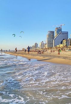 people are walking on the beach in front of some tall buildings and water with kites flying over them