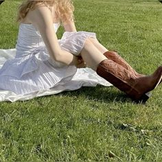 a woman sitting in the grass with her legs crossed and boots on, wearing a white dress