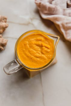 a glass cup filled with orange sauce on top of a white counter next to sliced ginger