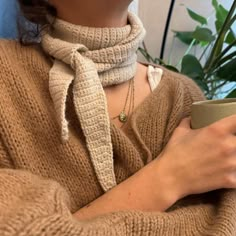 a woman is holding a coffee cup and wearing a scarf around her neck while sitting on a couch