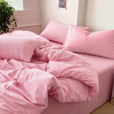 a bed with pink sheets and pillows in a room next to a potted plant