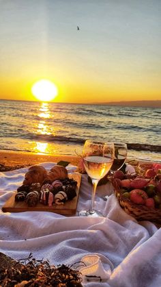 two glasses of wine and some food on a blanket at the beach with the sun setting in the background