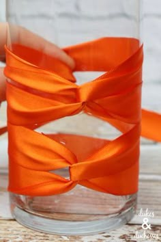 an orange ribbon wrapped around a clear glass vase with water in the bottom and one hand holding it