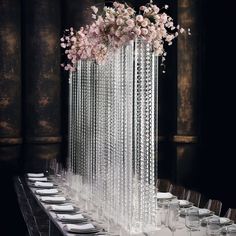 a long table with plates and wine glasses on it in front of a tall glass vase filled with flowers