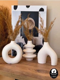 three white vases sitting on top of a wooden table with dried grass in them