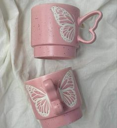 two pink mugs with white butterflies on them are sitting on a bed sheet and one has a heart shaped handle