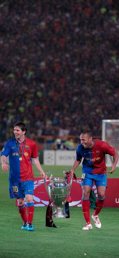 two soccer players are walking towards the trophy while another player is on the field behind them