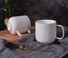 two white coffee mugs sitting on top of a wooden table next to a vase