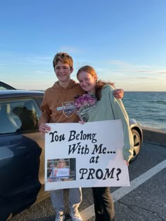 two people standing in front of a car holding a sign that says you belong with me at prom