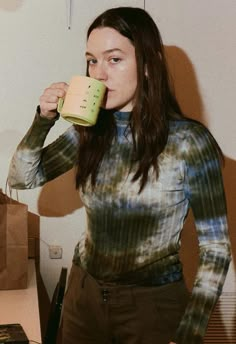 a woman drinking from a cup while standing in front of a desk