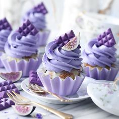 cupcakes with purple frosting and gold trim on plates next to utensils