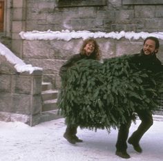 a man and woman walking in the snow carrying a christmas tree on their back,