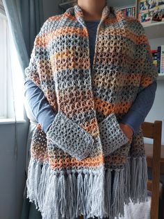 a woman standing in front of a window wearing a crocheted shawl with tassels