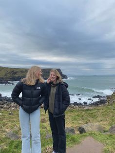 two women standing next to each other in front of the ocean on a cloudy day