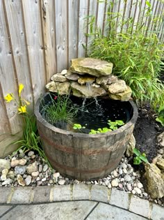 a wooden barrel filled with water next to rocks and flowers in a garden area near a fence