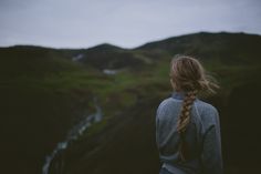 a woman with long hair standing in front of a mountain and looking at the water