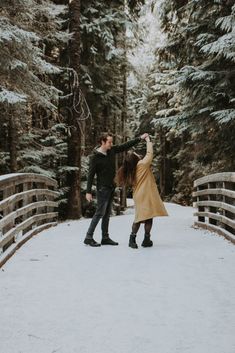 a man and woman standing on a bridge in the snow with trees behind them holding hands