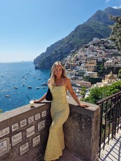 a woman in a yellow dress leaning on a wall near the ocean with boats and mountains in the background