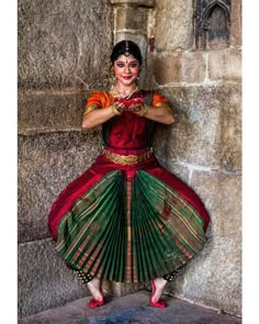 a woman in a red and green sari is standing by a stone wall with her hands on her hips