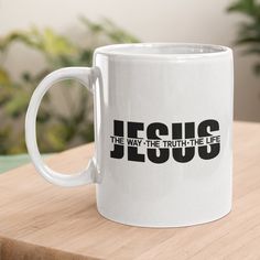 a white coffee mug with the word jesus on it sitting on a wooden table next to a potted plant