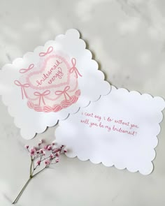 two cards with pink writing on them sitting next to a small purple flower in a vase