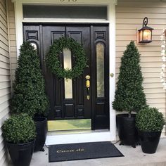 three potted plants on the front steps of a house with a black door and wreath