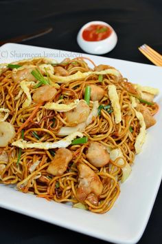 a white plate topped with noodles and shrimp on top of a black table next to a bowl of sauce