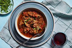 a bowl of stew with meat and vegetables on a blue table cloth next to a glass of red wine