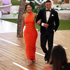 a man in a tuxedo and a woman in an orange dress walking down a wooden floor