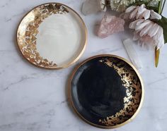 two black and gold plates sitting on top of a white table next to pink flowers