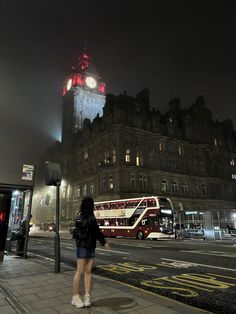 a woman standing on the sidewalk in front of a building with a clock tower at night