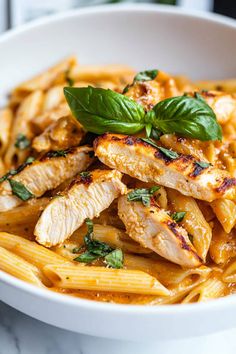 a white bowl filled with pasta and chicken covered in sauce, garnished with fresh basil