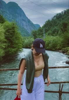 a woman standing on the side of a bridge next to a river with mountains in the background