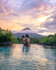 two people are sitting in the water at sunset with a mountain in the distance behind them