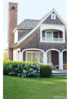 a house that is in the grass with flowers on the front lawn and bushes around it