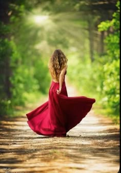 a woman in a red dress walking down a dirt road with her back to the camera