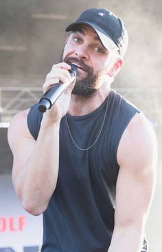 a man with a beard holding a microphone in his right hand and wearing a baseball cap