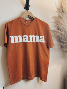 "Introducing the Mama T-shirt, exclusively available on Powered by Daisies! Celebrate motherhood in style with this comfortable and chic rust orange t-shirt. Made from high-quality materials, this t-shirt features the word \"Mama\" in bold letters. It's the perfect gift for yourself or any mom in your life - whether you're a new mom, a seasoned pro, or somewhere in between. Available in a range of sizes, our Mama T-shirt is designed to fit all body types comfortably. It's perfect for running err Graphic Tee With Letter Print As Gift, Letter Print Crew Neck Tops For Gifts, Graphic Tee Shirt With Name Print In Relaxed Fit, Orange Top With Letter Print And Relaxed Fit, Unisex Tops With Letter Print For Everyday, Family Matching Cotton T-shirt Gift, Custom Print Short Sleeve Tops For Everyday, Family Matching Crew Neck Cotton Tops, Cotton Tops With Name Print And Relaxed Fit