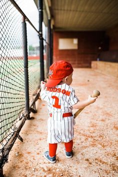 a young boy holding a baseball bat on top of a dirt field next to a fence