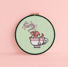 a cross - stitch picture of a teacup with a pink flower in it on a light green background