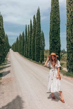 a woman in a dress and hat standing on a dirt road surrounded by tall trees