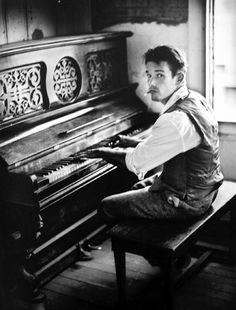 a man sitting on top of a wooden bench next to a piano in front of a window