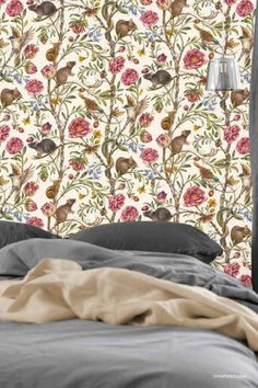 A bedroom with a wall featuring a detailed, vintage-style floral wallpaper pattern. The design includes branches, birds, and pink roses against a cream background. The bed in the foreground has dark gray pillows and blankets, with a beige throw draped across it. A modern silver lamp hangs on the right side near the bed. Wallpaper Of Flowers, William Morris Daisy Wallpaper, Birds And Thistle Wallpaper, Coloured Wallpaper, Pink Bird Toilet Wallpaper, William Morris Bird And Pomegranate Wallpaper, Flowers And Animals, Morris & Co. Bird And Pomegranate Wallpaper, Colourful Wallpaper