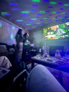 two people standing in a living room with lights on the ceiling and television above them