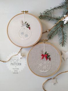 two cross stitch christmas ornaments hanging from twine hoops on a white background with pine branches