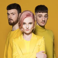 two men and a woman standing next to each other in front of a yellow background